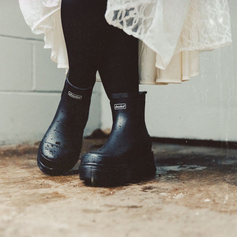 Black rubber rain boots with a white 'shaker' logo on the side, worn by a person in black tights and a white lace dress, standing on a wet concrete floor.