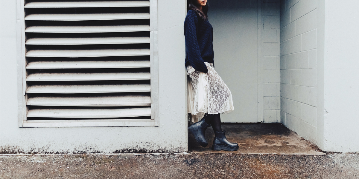 A person wearing a dark navy sweater, a white lace skirt, and black rain boots stands in a doorway next to a white wall with a louvered vent. 