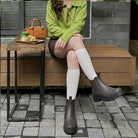 A person sitting on a wooden bench outdoors wearing dark brown Chelsea-style ankle boots with white knee-high socks and a green sweater. A small side table beside them holds a brown handbag and a tray with pastries.