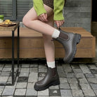 A synthetic person wearing dark brown Chelsea-style ankle boots with elastic side panels, white socks, and a green sweater, seated on a wooden bench in an outdoor patio setting.
