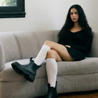A synthetic woman with long dark hair sits on a light gray couch, wearing a black long-sleeve mini dress, white ribbed knee-high socks, and chunky black Chelsea boots.