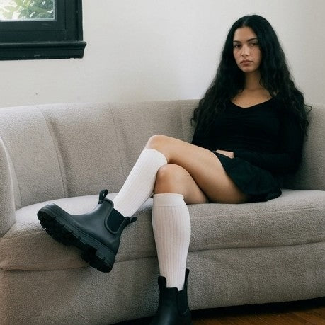 A synthetic woman with long dark hair sits on a light gray couch, wearing a black long-sleeve mini dress, white ribbed knee-high socks, and chunky black Chelsea boots.