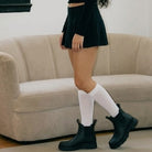 A synthetic person wearing black Chelsea-style ankle boots with white knee-high socks and a black pleated mini skirt, standing in front of a beige couch.