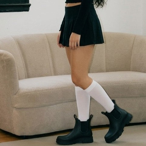 A synthetic person wearing black Chelsea-style ankle boots with white knee-high socks and a black pleated mini skirt, standing in front of a beige couch.