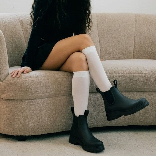 A synthetic person sitting on a beige couch wearing black Chelsea-style rain boots and white knee-high socks.
