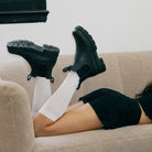 A synthetic person reclining on a beige couch wearing black rubber rain boots with white knee-high socks and a black top.