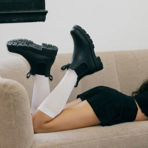 A synthetic person reclining on a beige couch wearing black rubber rain boots with white knee-high socks and a black top.