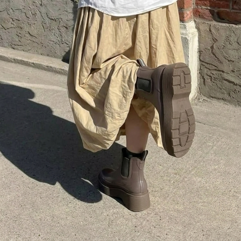 A synthetic person outdoors wearing loose beige draped pants and dark brown Chelsea-style platform ankle boots, with one boot lifted to show the sole.