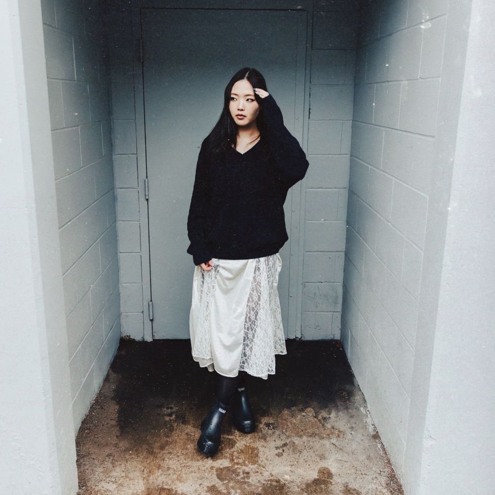 A woman stands in a narrow concrete corridor wearing a black oversized sweater, a white lace-trimmed midi skirt, black boots, and black socks.
