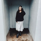 A woman stands in a narrow concrete corridor wearing a black oversized sweater, a white lace-trimmed midi skirt, black boots, and black socks.