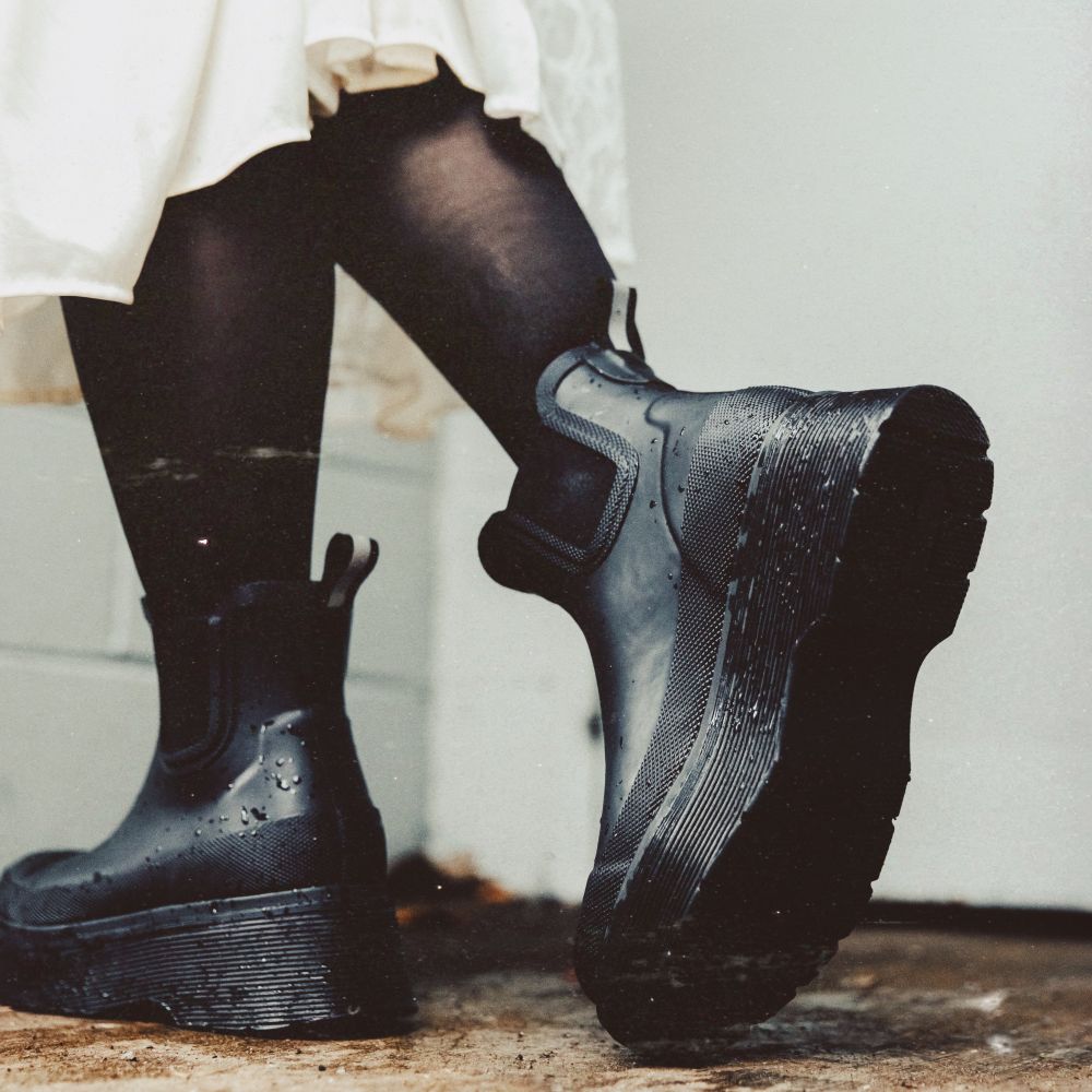 Black rubber Chelsea-style ankle boots with chunky lug soles, worn with black tights and a white skirt, photographed against a light wall.