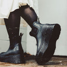 Black rubber Chelsea-style ankle boots with chunky lug soles, worn with black tights and a white skirt, photographed against a light wall.