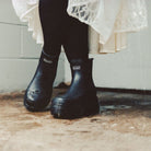Black rubber rain boots with a white 'shaker' logo on the side, worn by a person in black tights and a white lace dress, standing on a wet concrete floor.