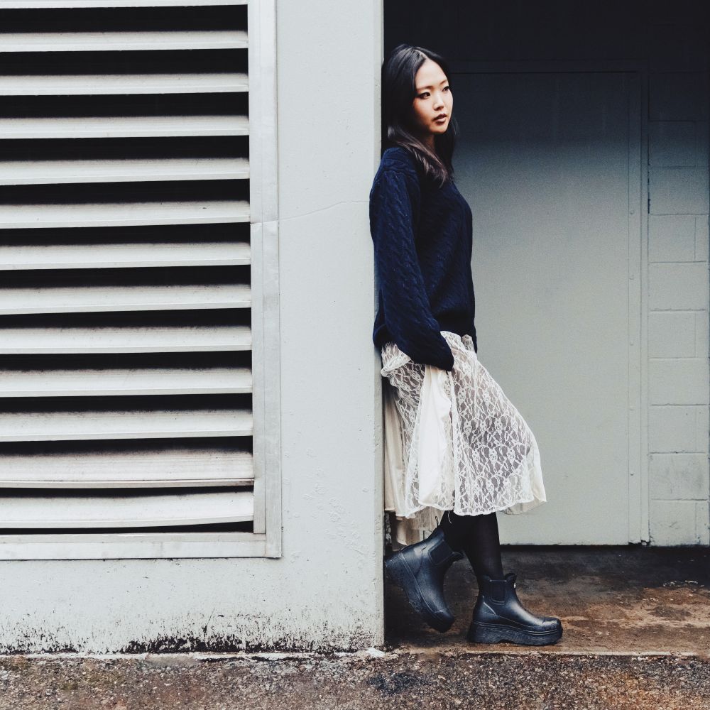 A woman stands in a doorway wearing a dark navy cable-knit sweater, a cream lace skirt, black tights, and black rubber boots, posed next to a white wall with horizontal louvers.