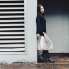 A woman stands in a doorway wearing a dark navy cable-knit sweater, a cream lace skirt, black tights, and black rubber boots, posed next to a white wall with horizontal louvers.